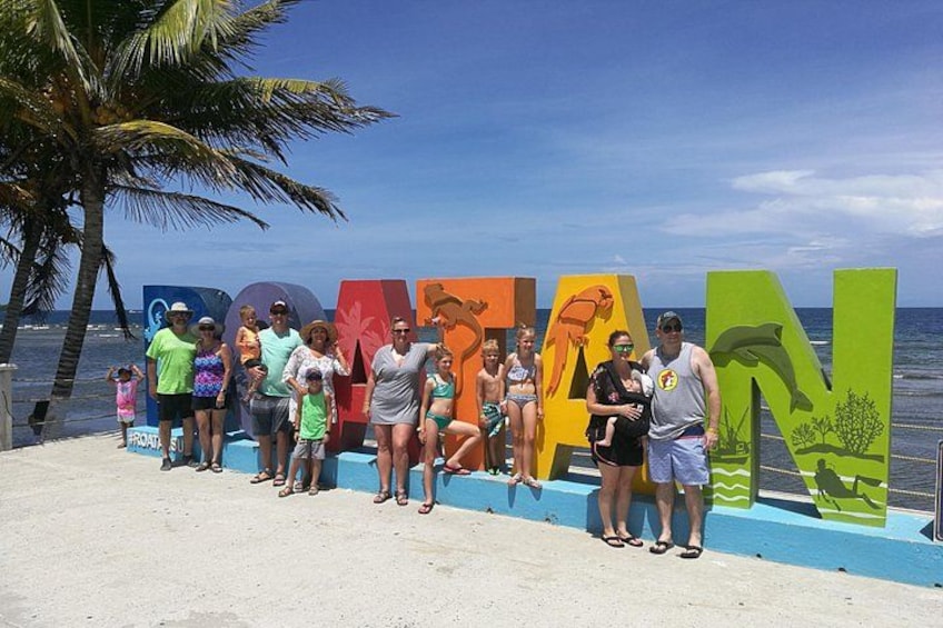 A group of people standing in front of colorful letters spelling 'BANANAN' on a beach with palm trees and the ocean in the background. 1