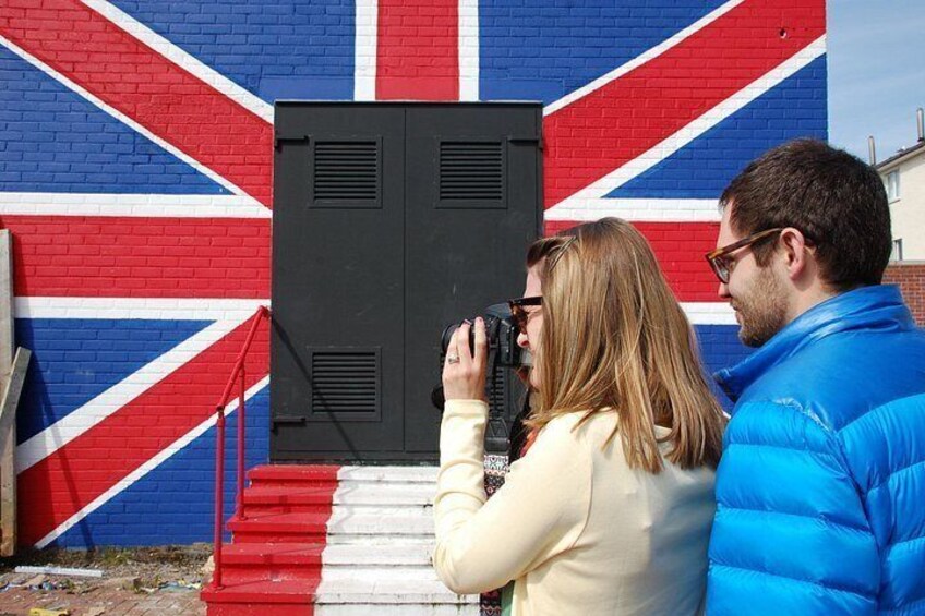 A person taking a photo with a camera in front of a wall with a Union Jack flag design. 4