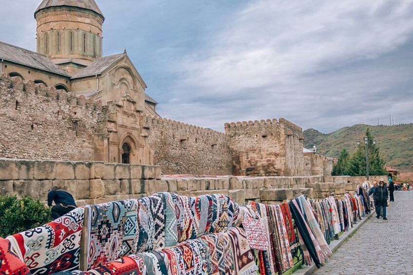 Un muro de piedra histórico con una iglesia y una torre, textiles coloridos exhibidos, y un camino adoquinado. 2