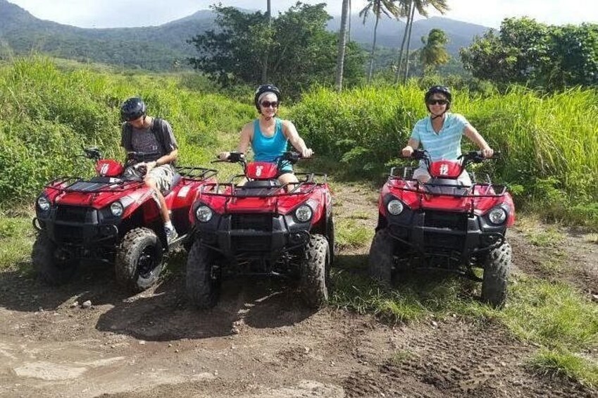 Three people on red ATVs in a rural, grassy area with mountains in the background. 1