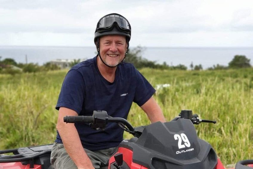 Une personne est assise sur un quad rouge portant le numéro 29, elle porte un casque et un t-shirt bleu foncé, et se trouve dans un champ herbeux avec une étendue d'eau en arrière-plan. 3