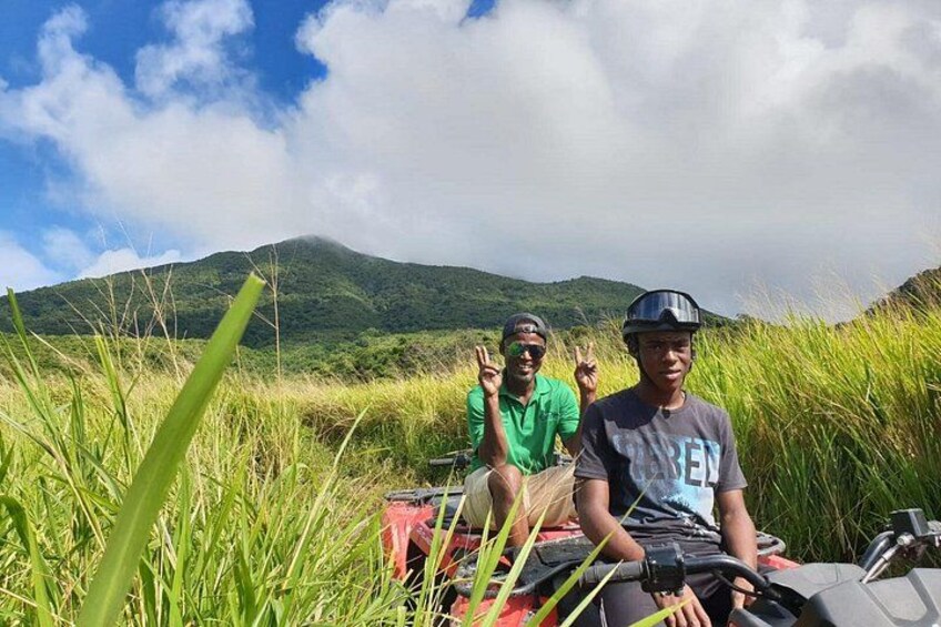 Two people sitting on a red vehicle in a grassy field with mountains in the background. 5