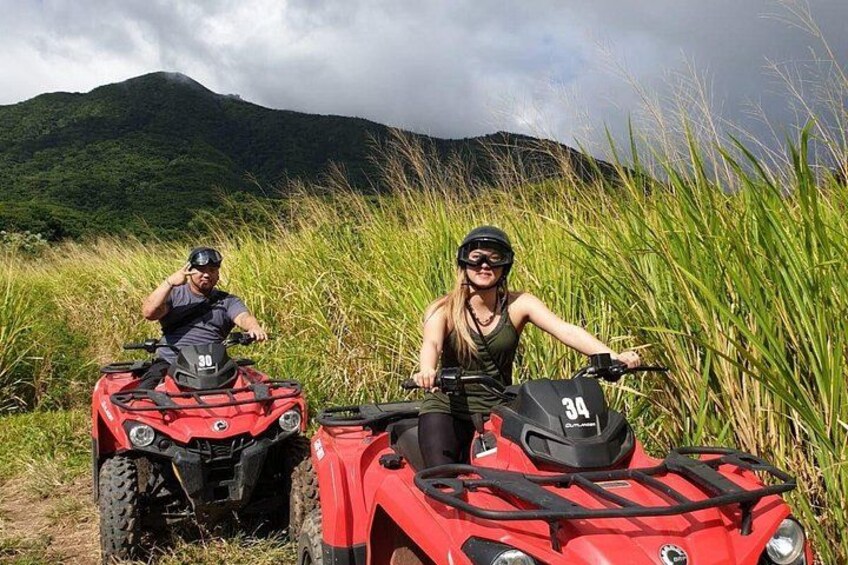 Two people on red ATVs in a grassy field with mountains in the background. 2