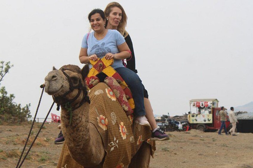 Two people riding a camel in a desert setting with a vehicle in the background. 3