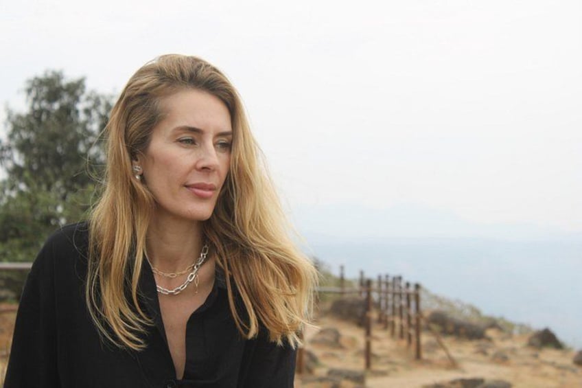 A person with blonde hair wearing a black top and a necklace, standing outdoors with a coastal landscape in the background. 5