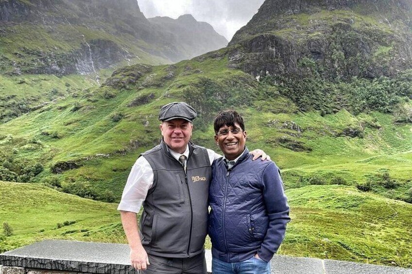 Two people standing on a ledge with a mountainous landscape in the background. 1