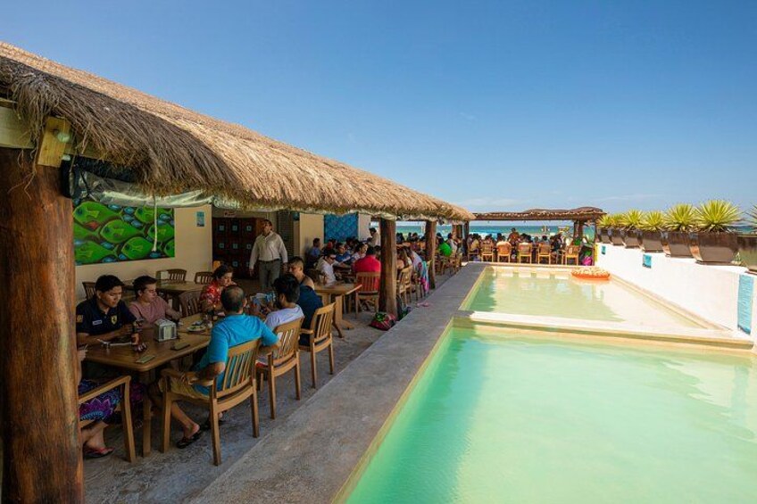 Un área de comedor junto a la piscina con mesas y sillas de madera, una estructura con techo de paja y un cielo azul despejado. 4