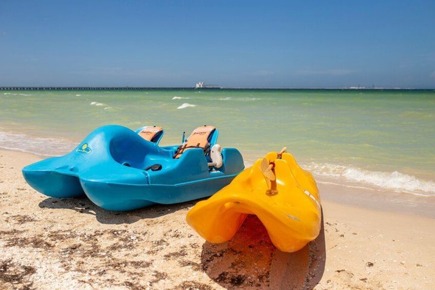 Deux kayaks de mer bleus et un jaune sont posés sur une plage de sable, avec un ciel bleu dégagé et une mer calme en arrière-plan. 2