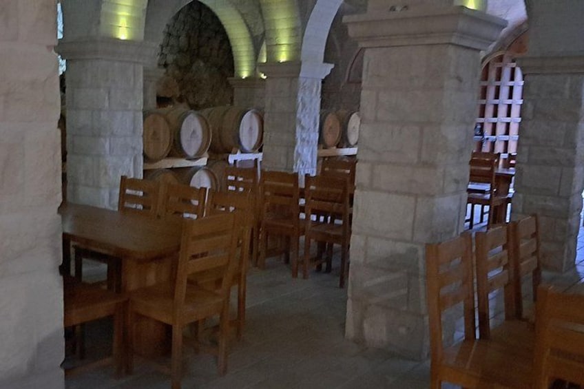 Una habitación de paredes de piedra con mesas y sillas de madera, barriles y una chimenea de piedra. 3