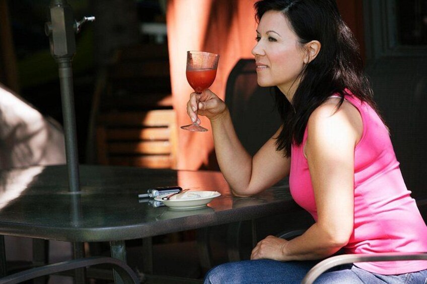 A person sitting at an outdoor table with a glass of red beverage, a plate, and cutlery. 2