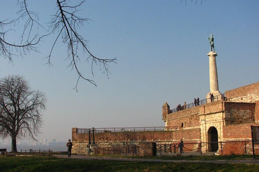 Un fort historique, surmonté d’une haute colonne et d’une statue, entouré d’un mur de briques et d’une clôture. 1