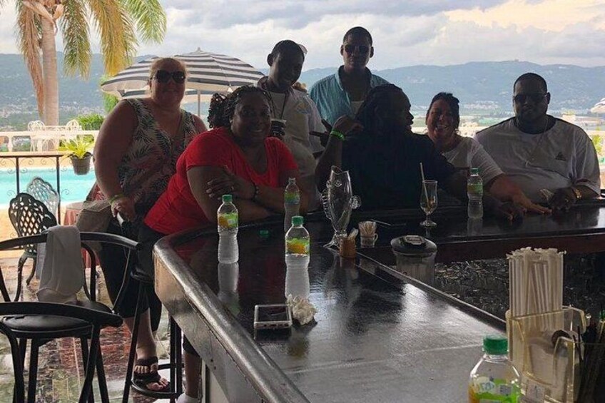 En grupp människor sitter vid en bar utomhus, med en pool och palmer i bakgrunden. 5
