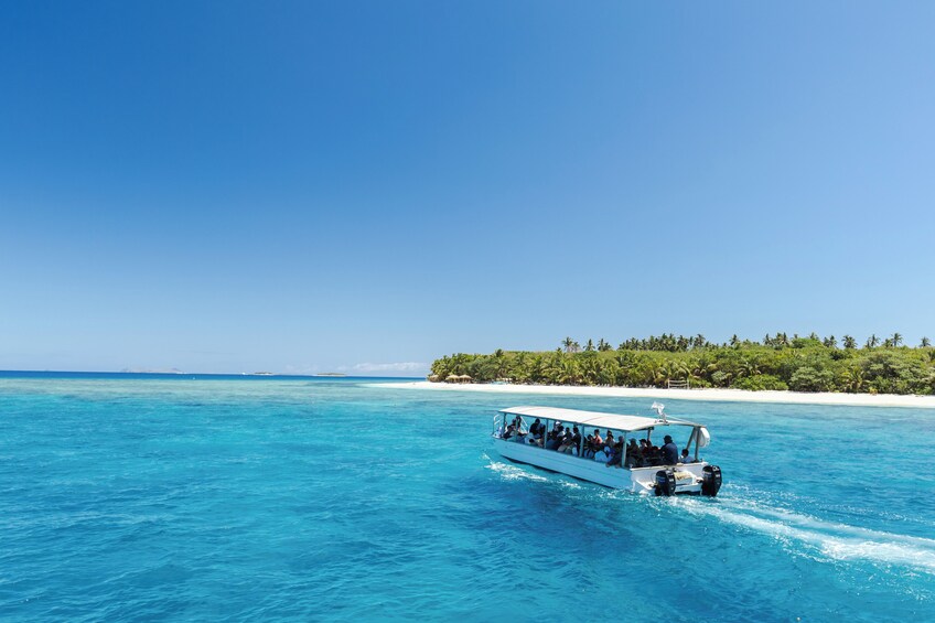 A boat with passengers on a clear blue sea, a sandy beach, and a forested island in the background. 1