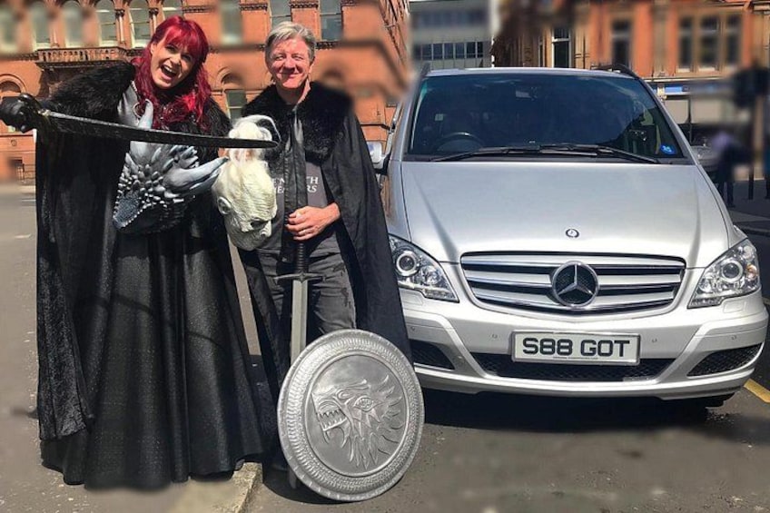 A silver car with a license plate, a person in a black cloak holding a white object, and a large coin on the ground. 3