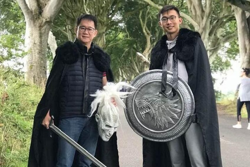 Two individuals in black coats holding a shield and a staff, standing in a park with trees. 5