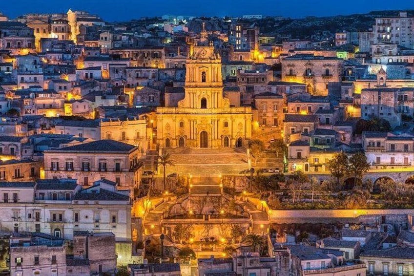 A cityscape at night with a prominent cathedral illuminated in the center, surrounded by densely packed buildings. 3