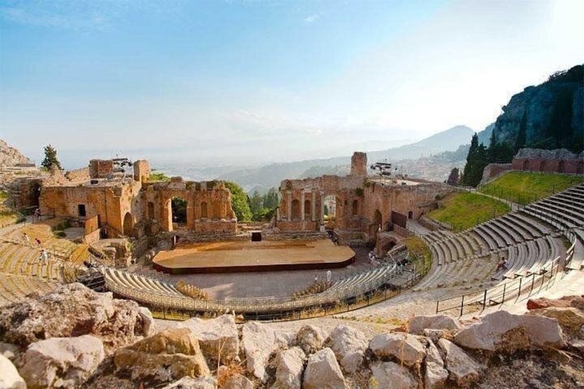 Un antico anfiteatro con rovine e un palcoscenico, circondato da terreno roccioso e montagne. 1