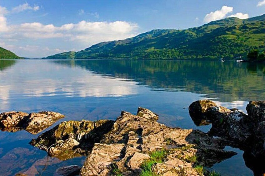 A calm lake with rocky shores, surrounded by green hills and a clear sky. 2