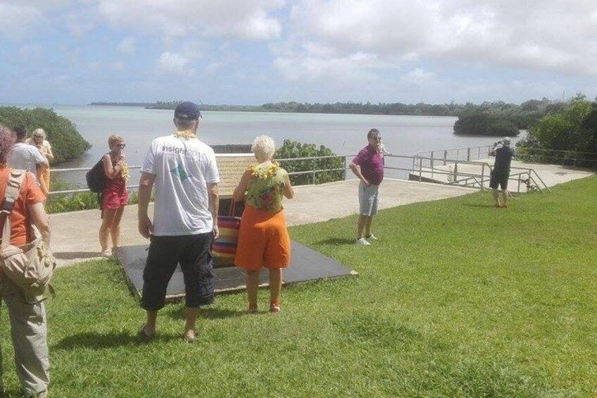 A group of people standing on a grassy area near a body of water, with a metal railing and a paved walkway in the background. 3
