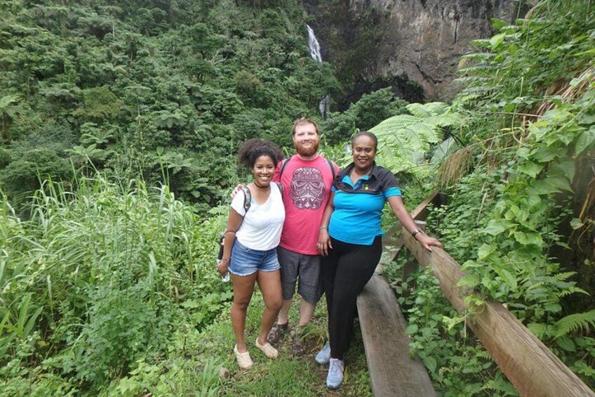 Three people standing on a wooden bridge surrounded by dense greenery and a waterfall in the background. 3