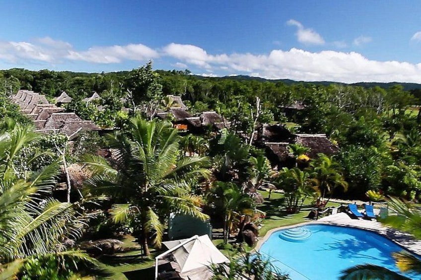 A tropical resort with a swimming pool, palm trees, and thatched-roof buildings. 2