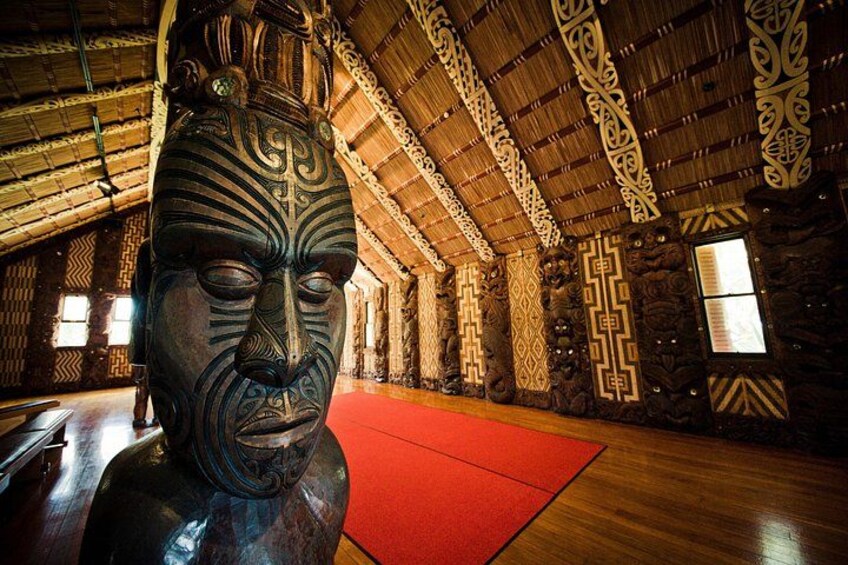 A wooden interior with a large carved wooden head sculpture, patterned walls, and a red mat on the floor. 3