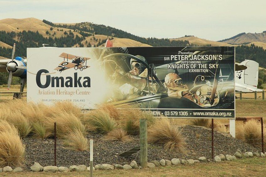 A sign for the Omaka Aviation Heritage Centre with a display of vintage aircraft and a mountainous landscape in the background. 2