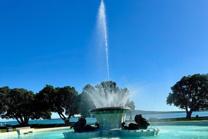Une fontaine avec des jets d’eau, entourée d’arbres et surplombée par un ciel bleu clair. 2
