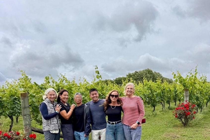 A group of people standing in a vineyard with rows of grapevines and red flowers in the background. 2
