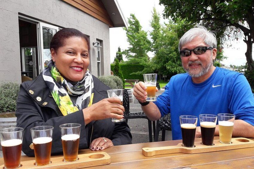 Two people sitting at a table with a wooden tray holding several glasses of beer. 5