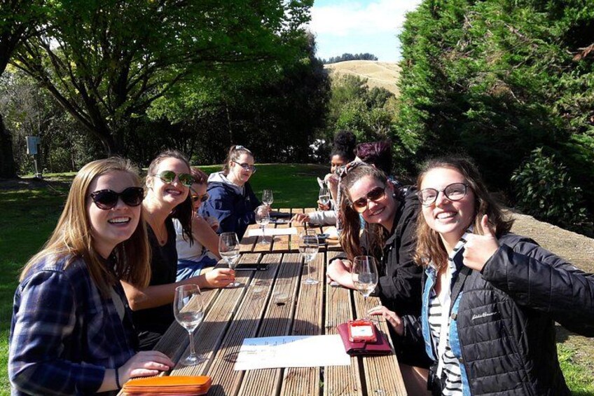 A group of people sitting at a wooden picnic table outdoors, with trees and hills in the background. 4