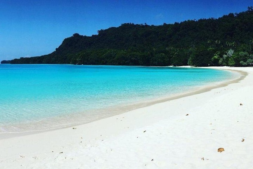 A beach with clear turquoise water, white sand, and a forested hill in the background. 5