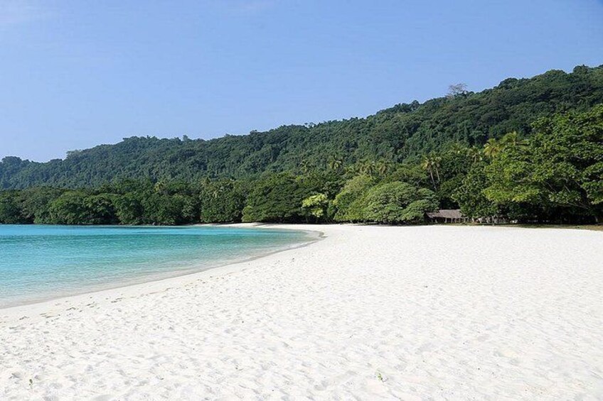 A white sandy beach with clear turquoise water and a forested hill in the background. 3