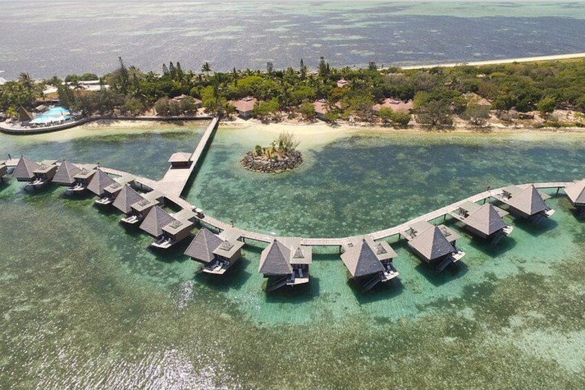 Vue aérienne de bungalows sur pilotis reliés par une jetée, entourés d’eaux turquoise cristallines et d’un paysage tropical. 2