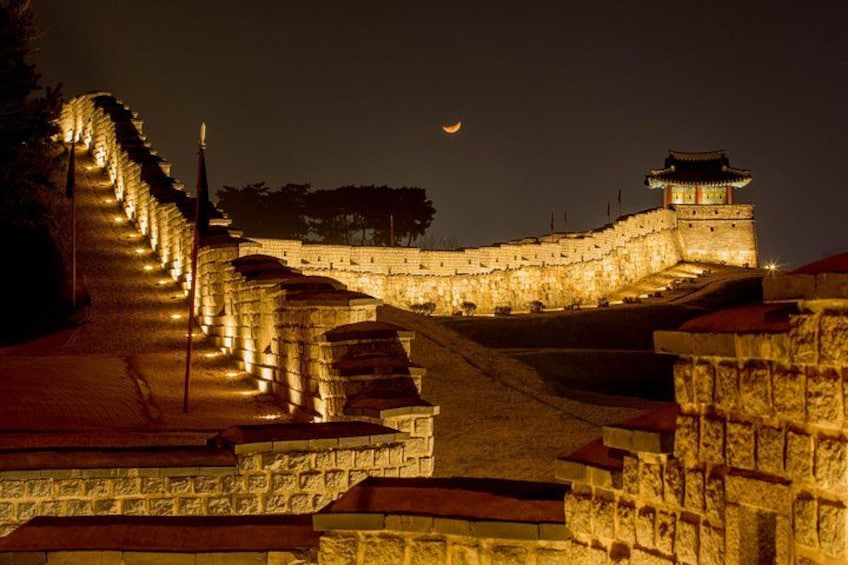 Un mur de pierre surmonté d’une tour de guet, éclairé par un système d’éclairage artificiel, sous un ciel nocturne où l’on aperçoit la lune. 4