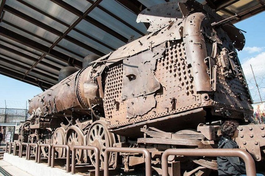 A large, rusted locomotive with visible wear and tear, including dents and scratches, parked under a metal roof structure. 5