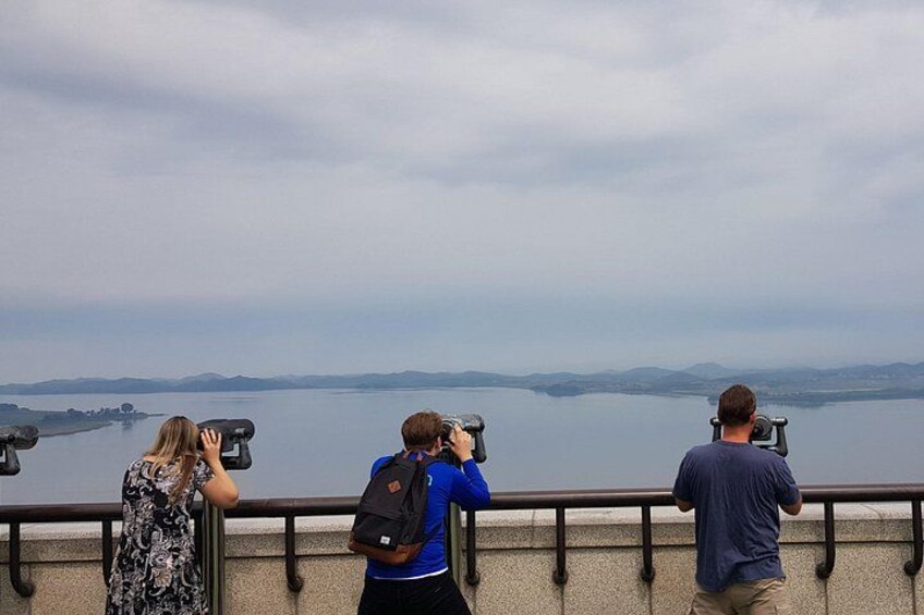 Three people using binoculars to observe a body of water and distant land. 1