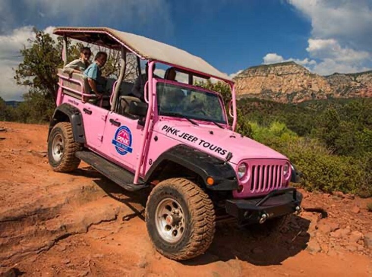 Un Jeep rose, sur lequel on peut lire « PINK JEEP TOURS », est garé sur un terrain rocailleux, avec un ciel dégagé et des montagnes en arrière-plan. 3