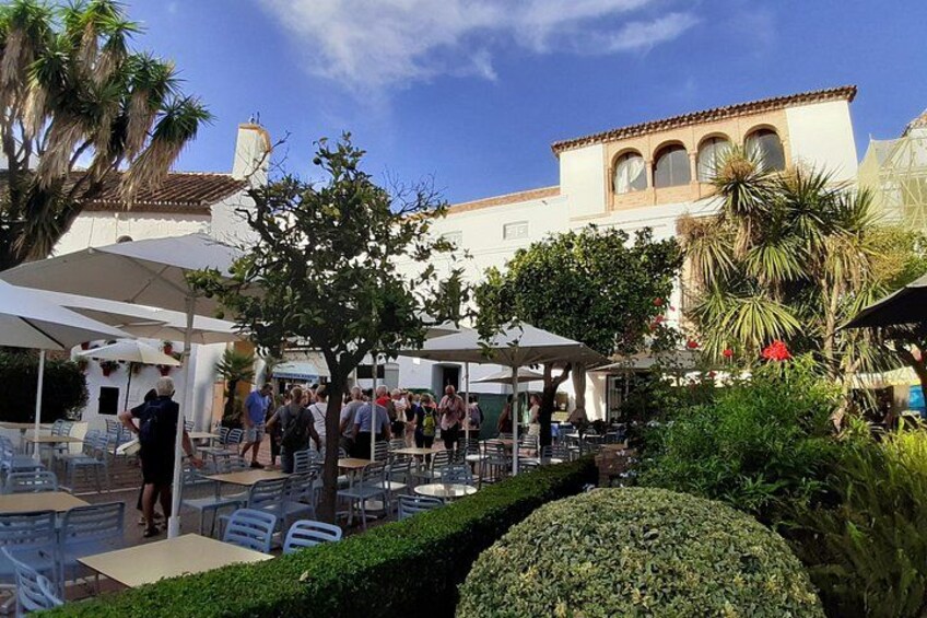 Un café al aire libre con mesas y sillas, rodeado de follaje y un edificio con tejado de azulejos. 1