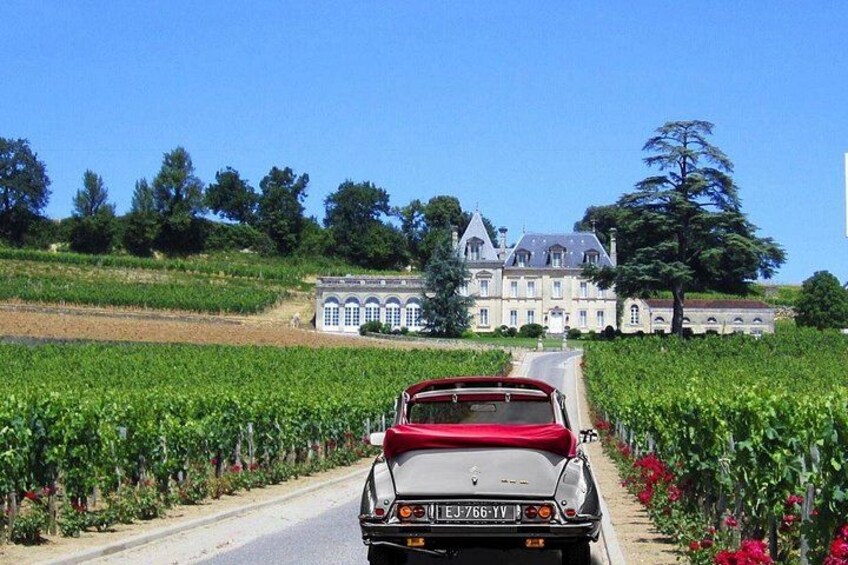 Une voiture ancienne roule sur une route, avec en arrière-plan un vaste domaine et un vignoble. 1