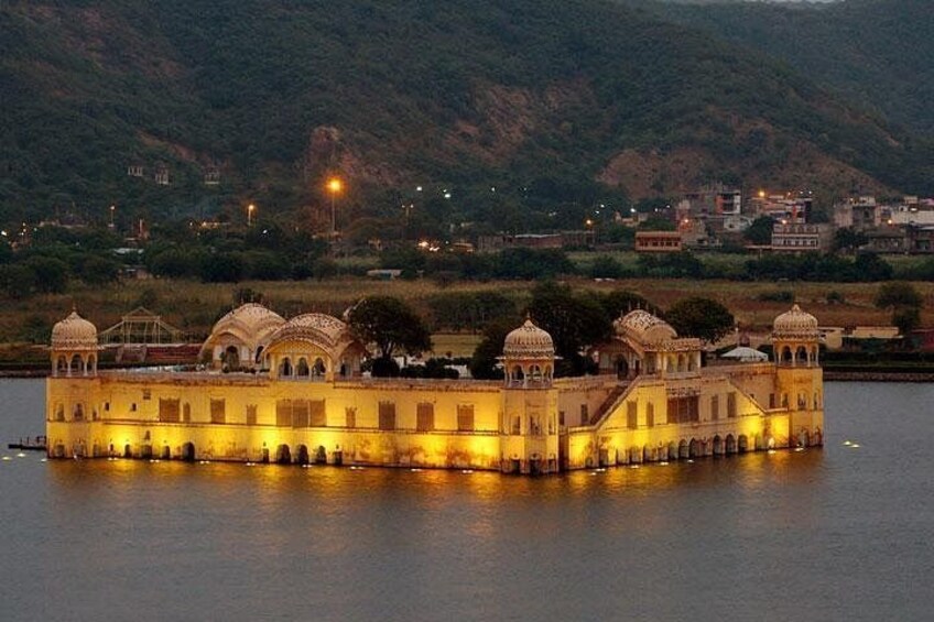 Un palazzo con strutture a cupola su un lago, circondato da colline e luci cittadine sullo sfondo. 1