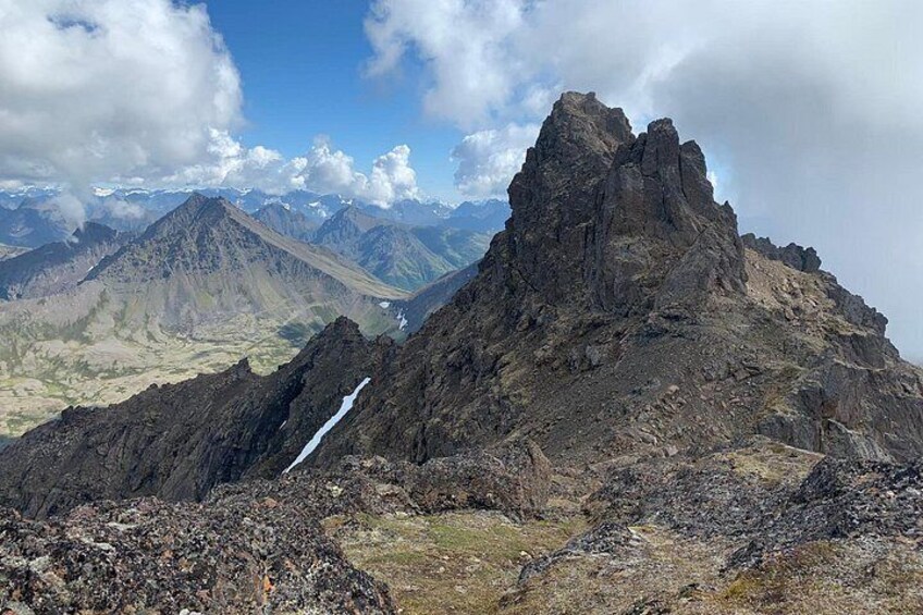 A rugged mountain peak with a glacier, a valley, and distant mountains under a partly cloudy sky. 2
