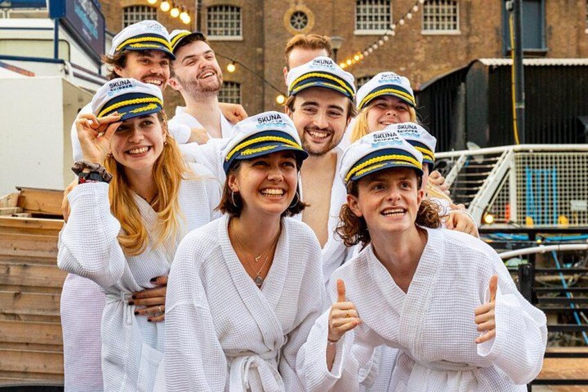 A group of people wearing white robes and matching hats with a logo, standing in front of a brick building with a staircase and string lights. 4