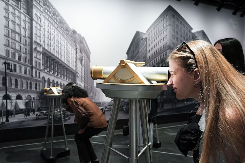 Eine Person betrachtet mit einem Teleskop eine Museumsausstellung mit historischen Stadtansichten an der Wand. 1