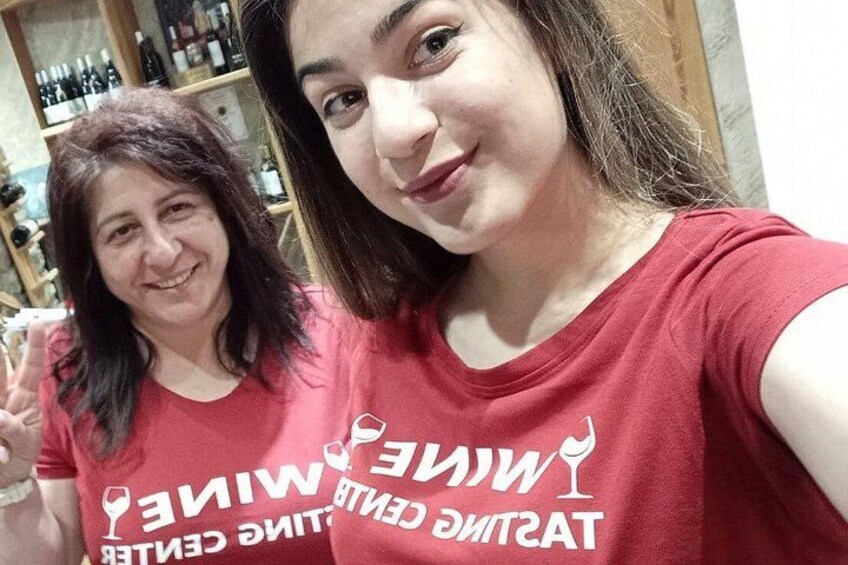 Two people wearing red t-shirts with white text, standing indoors with a shelf of bottles in the background. 5