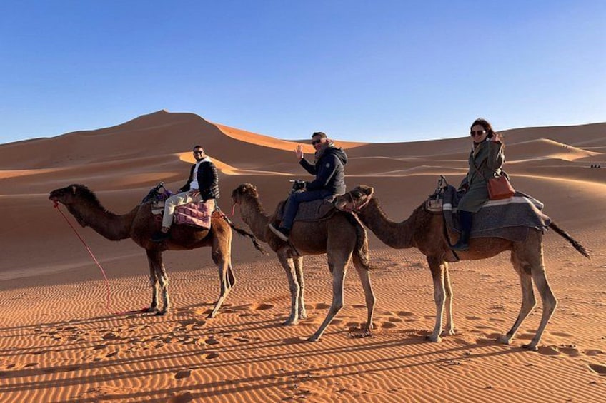 Three people riding camels in a desert with sand dunes in the background. 4
