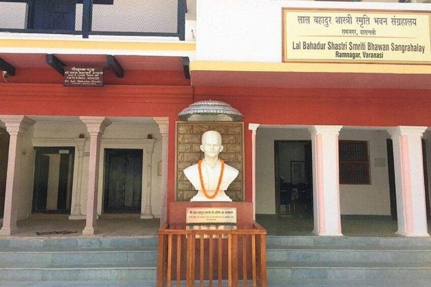 A memorial with a bust and a plaque in front of a building with columns. 1