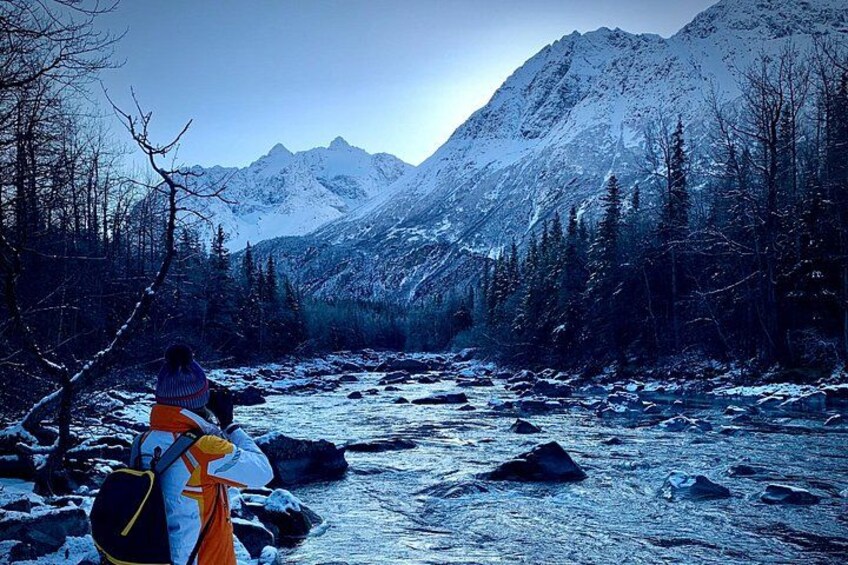 A person standing by a snow-covered river with mountains in the background. 5