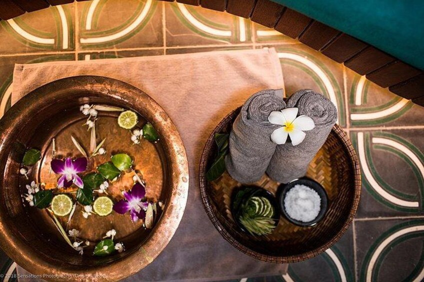 A spa setting with a copper bowl containing water, flowers, and lime slices. There are rolled towels and a flower on a wooden tray. 3