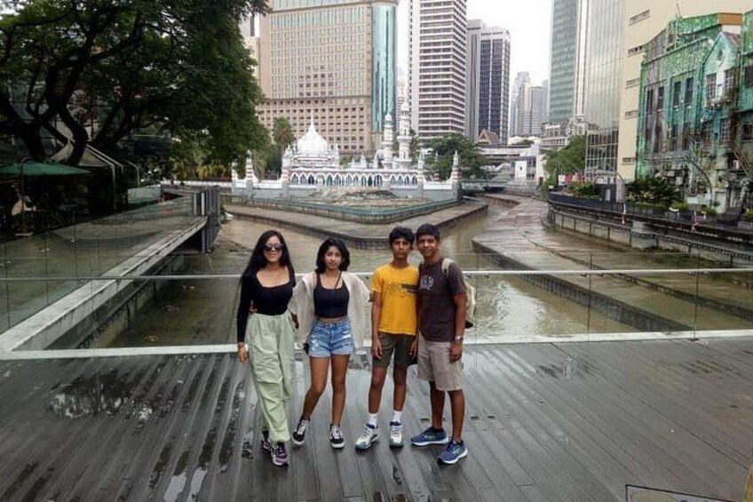 A group of people standing on a wooden boardwalk with a cityscape and a water feature in the background. 2
